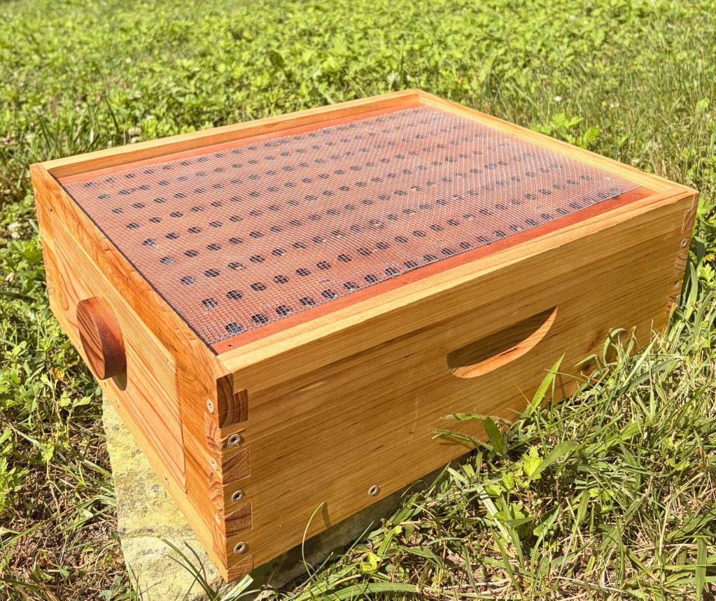 An outdoor close-up of the wooden ThermaHive V2 bottom treatment box resting on the grass with its solid blank insert installed